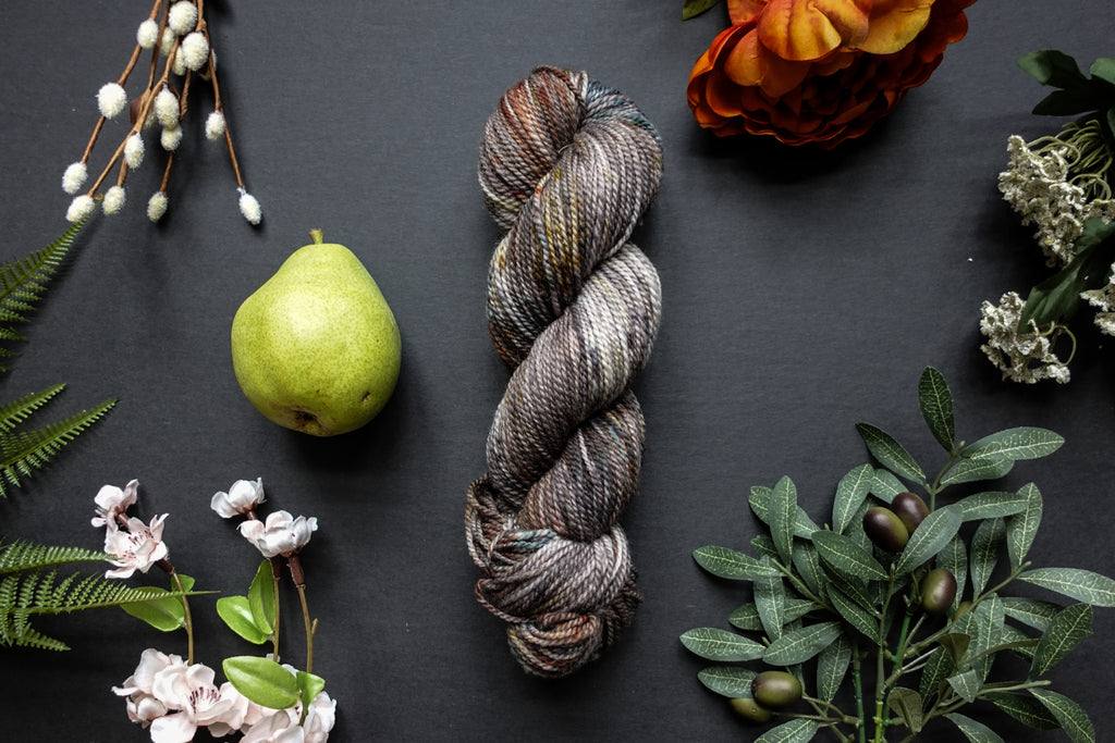 A variegated skein of brown, white, and beige DK weight yarn lies on a black surface. It's surrounded by flowers, branches, an orange rose, and a pear.