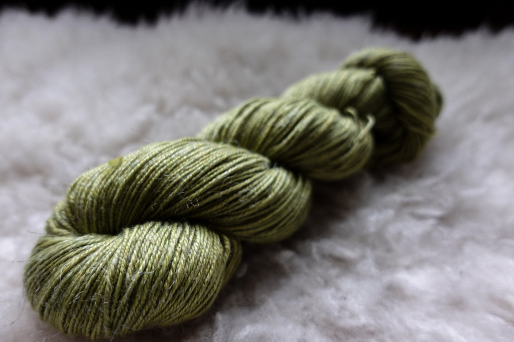 A skein of natural yarn has been hand dyed bright green. It lays on a sheepskin and is seen from the side.