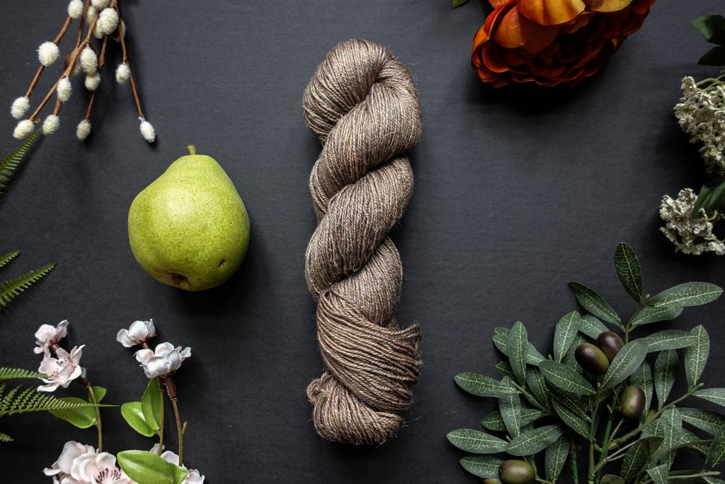 A beige brown skein of sport weight yarn lies on a black surface. It's surrounded by flowers, branches, an orange rose, and a pear.