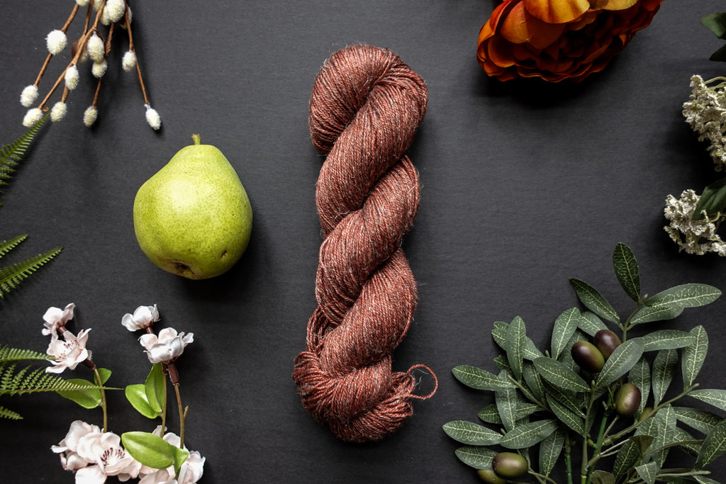A brick red skein of sport weight yarn lies on a black surface. It's surrounded by flowers, branches, an orange rose, and a pear.