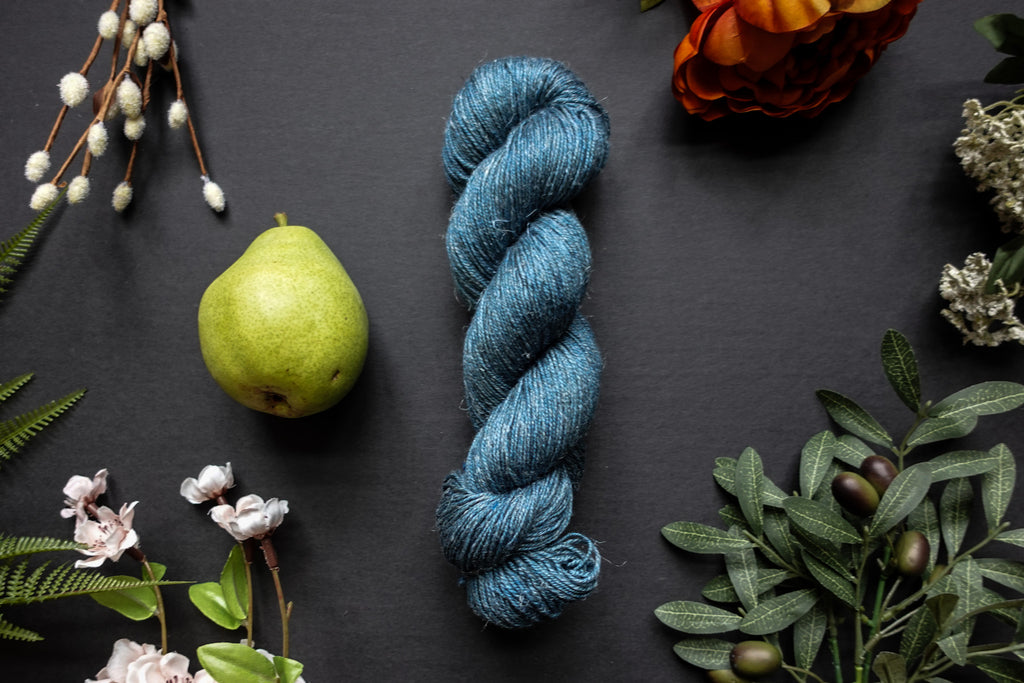 A skein of blue sport weight yarn lies on a black surface. It's surrounded by flowers, branches, an orange rose, and a pear.