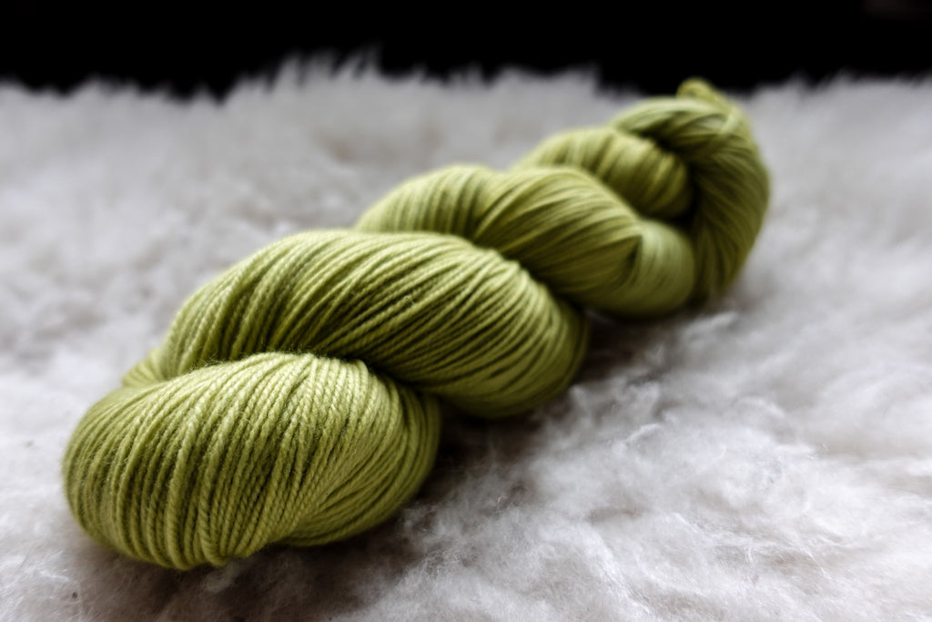 A skein of natural yarn has been hand dyed bright green. It lays on a sheepskin and is seen from the side.