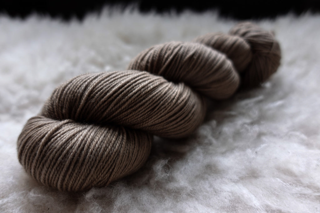 A natural skein of yarn has been hand dyed a light brown. It lays on a sheepskin and is seen from the side.