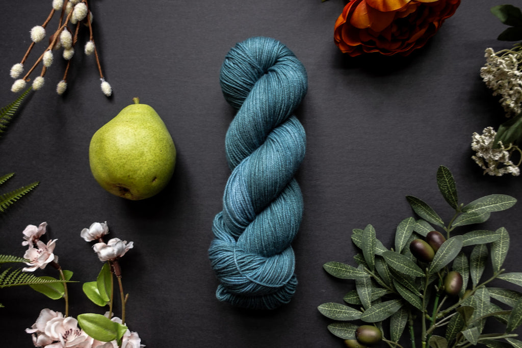 A skein of blue sock weight yarn lies on a black surface. It's surrounded by flowers, branches, an orange rose, and a pear.