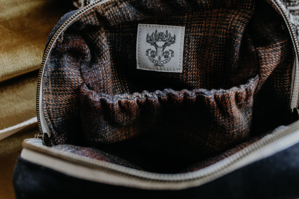 A black canvas yarn project bag is unzipped to show a brown plaid interior. There's an interior pouch, and the Forest Fiber Co. logo is sewn inside.
