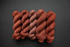 Five skeins of yarn, all different bases, are naturally dyed a brick red. They are lined up in a row on a black surface.