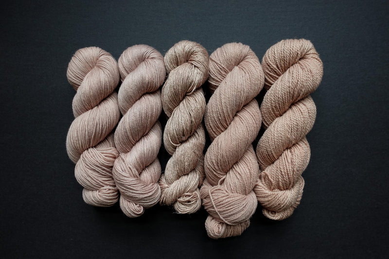 Five skeins of yarn, all different bases, are naturally dyed a light pink-beige. They are lined up in a row on a black surface.