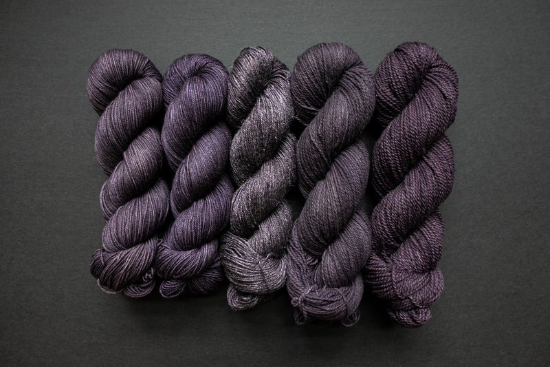 Five skeins of yarn, all different bases, are naturally dyed a deep purple. They are lined up in a row on a black surface.