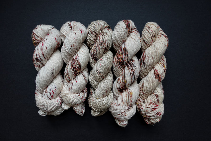 Five skeins of yarn, all different bases, are naturally dyed white with burgundy speckles. They are lined up in a row on a black surface.