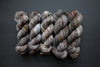 Five skeins of yarn, all different bases, are naturally dyed white, brown, and gray. They are lined up in a row on a black surface.