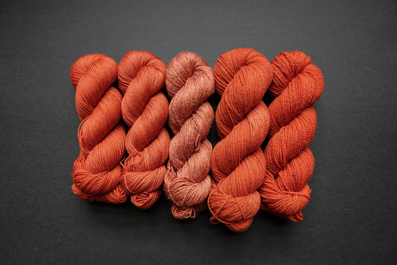 Five skeins of yarn, all different bases, are naturally dyed an orange-red. They are lined up in a row on a black surface.