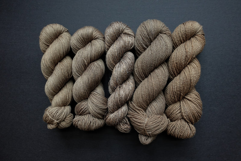 Five skeins of yarn, all different bases, are naturally dyed a light brown. They are lined up in a row on a black surface.