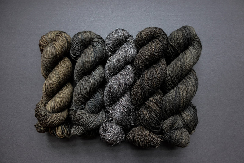 Five skeins of yarn, all different bases, are naturally dyed a dark muddy green, almost black. They are lined up in a row on a black surface.