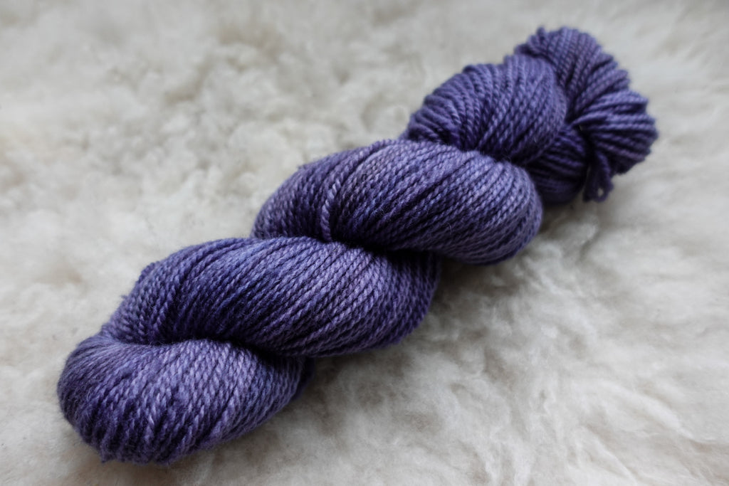 A hand dyed skein of purple yarn lays on a wool background.