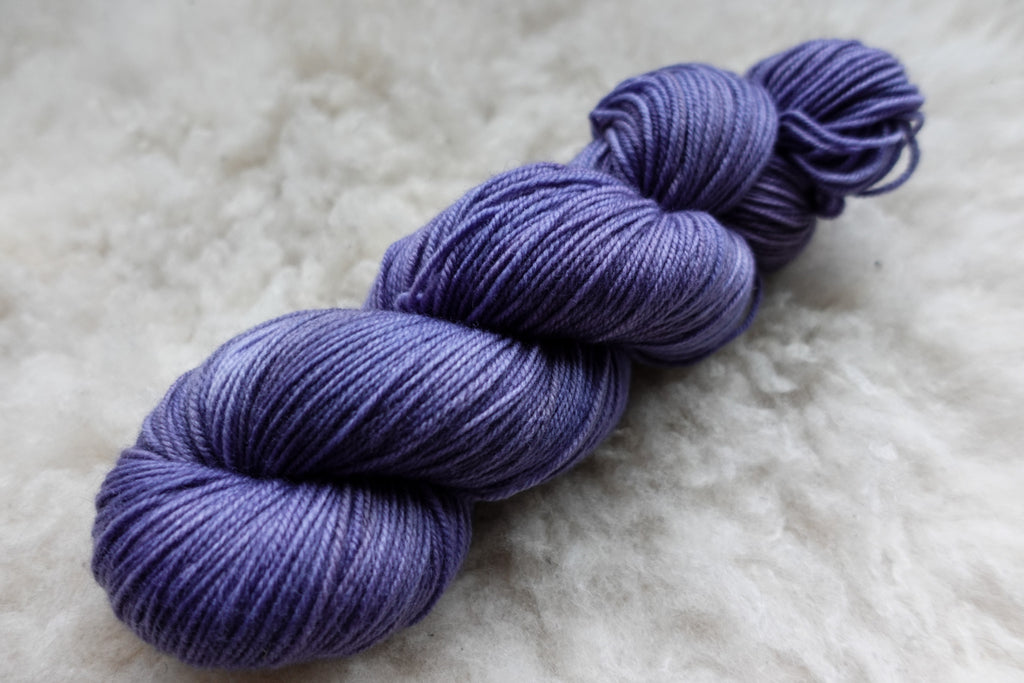 A fingering weight skein of yarn lays on a wool background. It has been hand dyed purple.