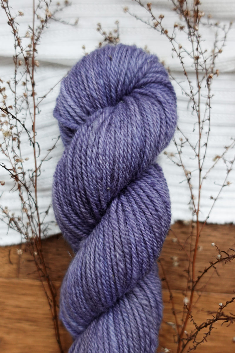 A skein of yarn has been naturally dyed purple. It lays on a table next to sprigs of budding twigs.