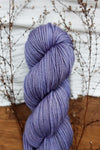 A natural fiber skein of yarn has been hand dyed purple. It lays on a tabletop next to budding twigs.