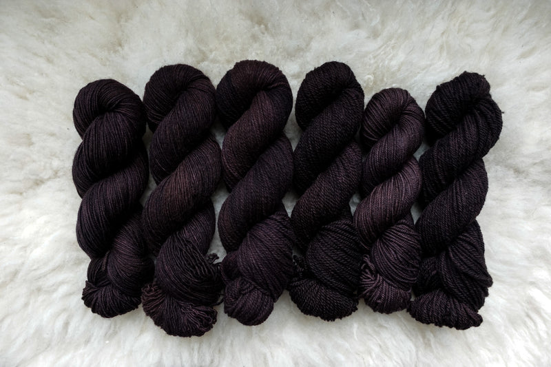 Six skeins of yarn, all different bases, have been hand dyed a dark burgundy. They lay on a sheepskin rug.