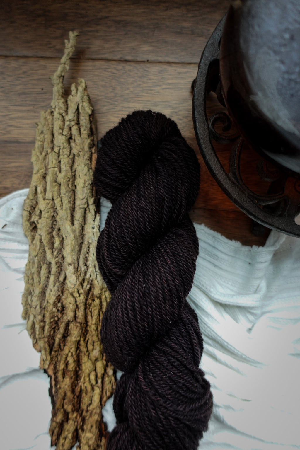 A skein of yarn, naturally dyed a dark burgundy, lays on a tabletop next to a strip of bark and a teapot.