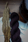 A hand dyed skein of dark burgundy yarn lays on a tabletop next to a strip of bark and a teapot.
