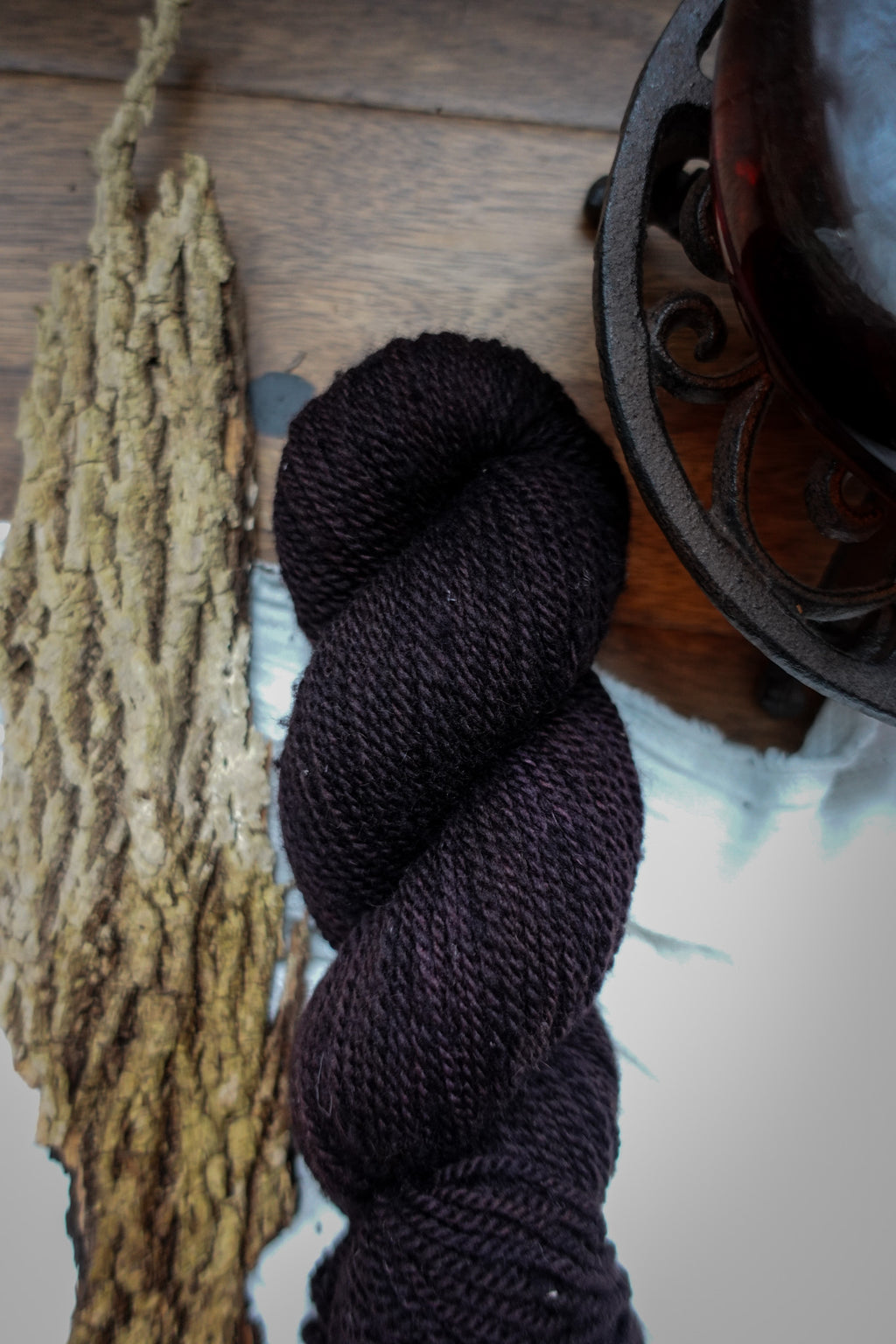 A skein of hand dyed, dark burgundy yarn lays on a tabletop next to some bark and a teapot.