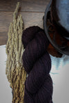 A dark burgundy skein of sport weight yarn lays on a tabletop, next to a strip of bark and a teapot.