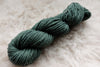 A skein of worsted weight, natural fiber yarn has been dyed blue green. It lays on a wool background.