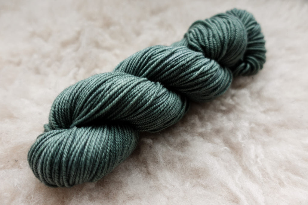 A skein of natural fiber yarn has been hand dyed a blue green. It lays on a wool background.