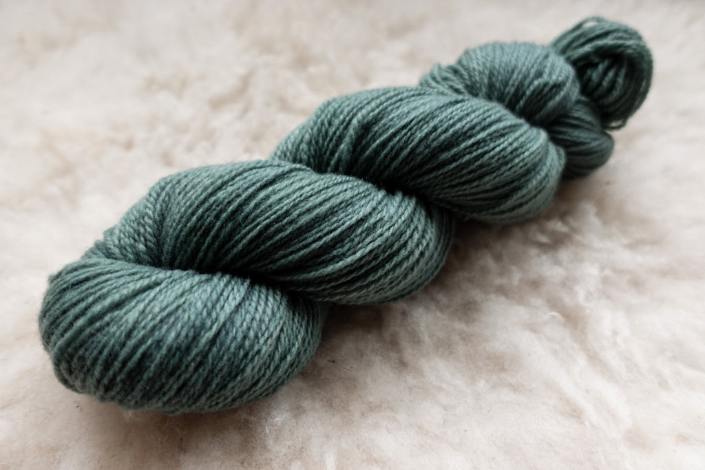 A skein of natural fiber yarn has been hand dyed blue green. It lays on a wool background.