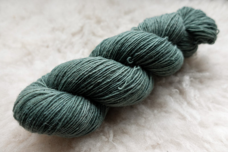 A single ply skein of fingering weight yarn has been hand dyed blue green. It lays on a wool background.