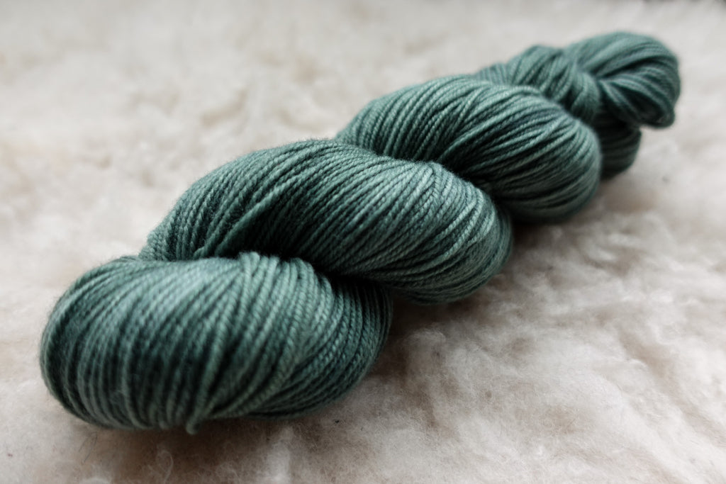 A skein of fingering weight yarn has been naturally dyed a blue spruce. It lays on a wool background.