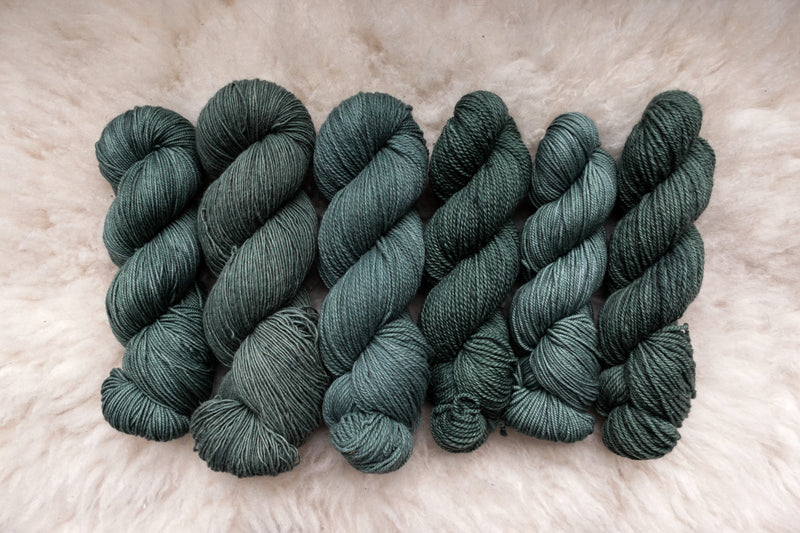 Six skeins of yarn, all different bases, have been hand dyed a blue-green. They lay on a sheepskin rug.