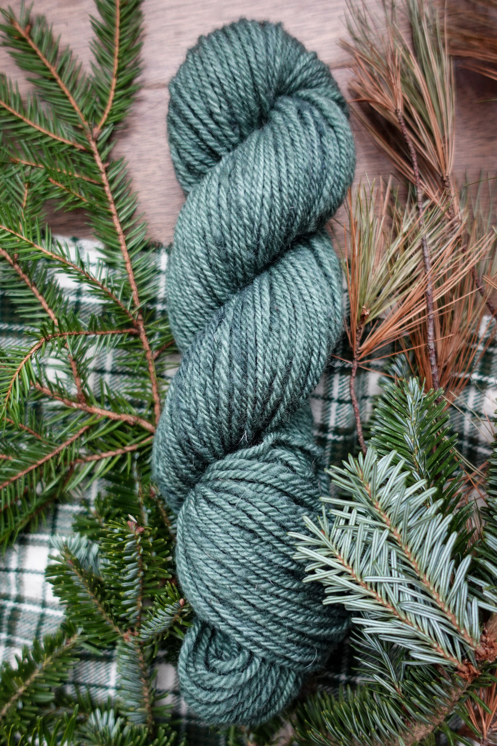 A skein of worsted weight yarn has been hand dyed blue green. It lays on a tabletop next to spruce branches.