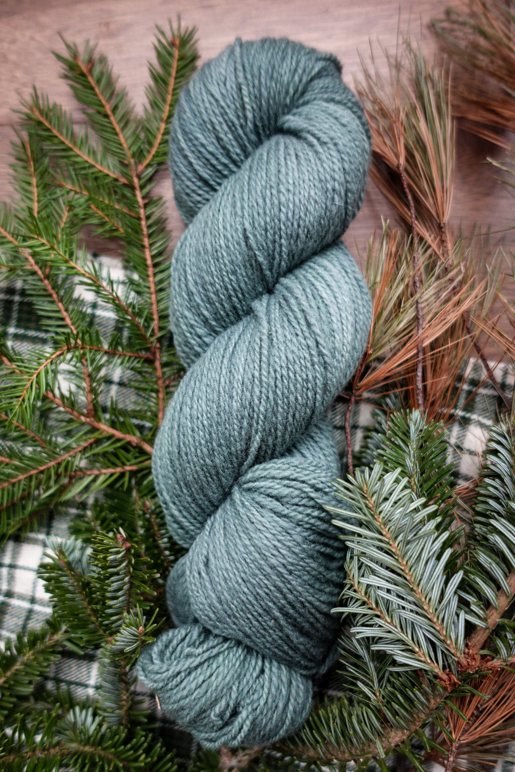 A skein of sport weight yarn has been hand dyed blue green. It lays on a tabletop next to spruce branches.