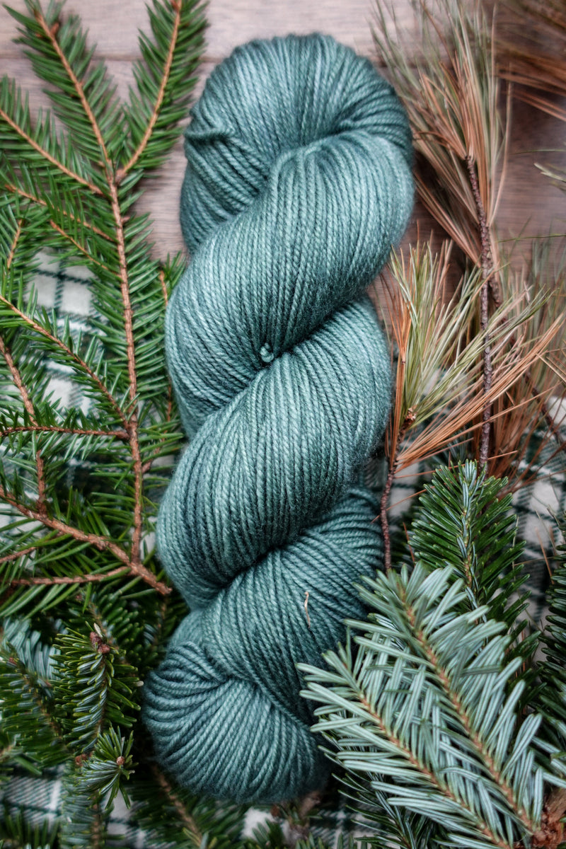 A naturally dyed, blue green skein of yarn lays on a tabletop next to spruce branches.