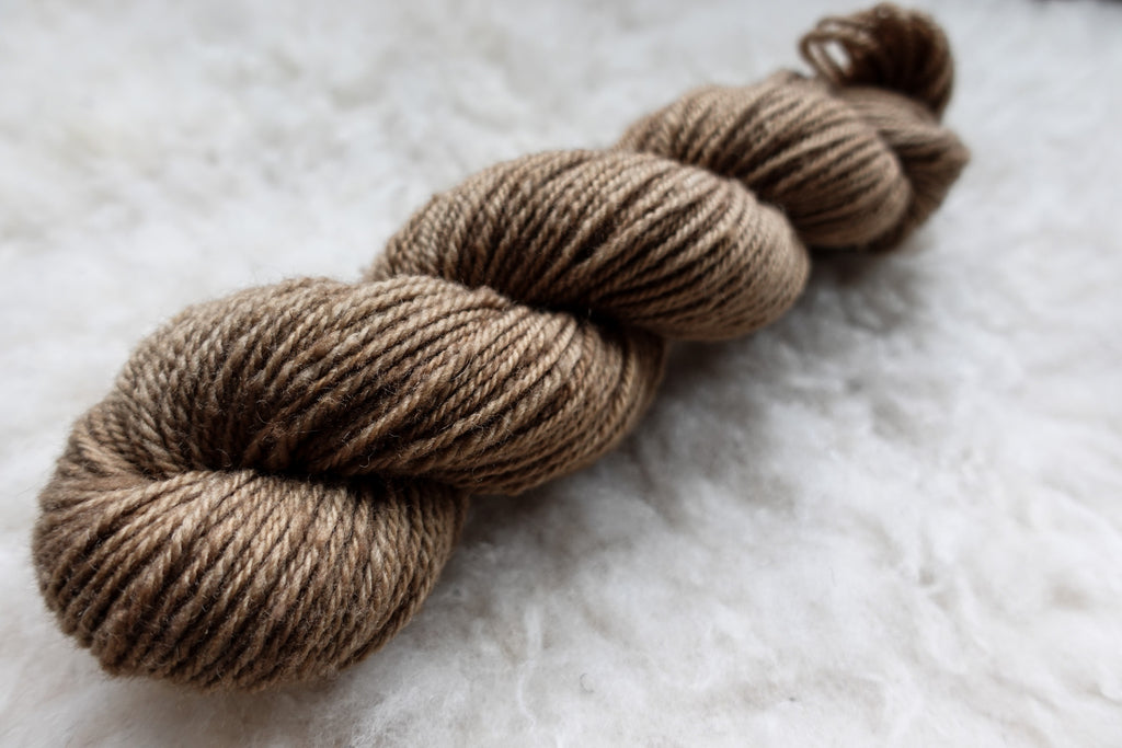 A worsted weight skein of natural fiber yarn has been hand dyed a light brown. It lays on a wool background.
