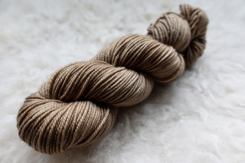 A worsted weight skein of natural fiber yarn has been hand dyed a light brown. It lays on a wool background.