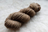 A DK weight skein of natural fiber yarn has been hand dyed a light brown. It lays on a wool background.