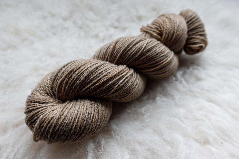 A sport weight skein of natural fiber yarn has been hand dyed a light brown. It lays on a wool background.