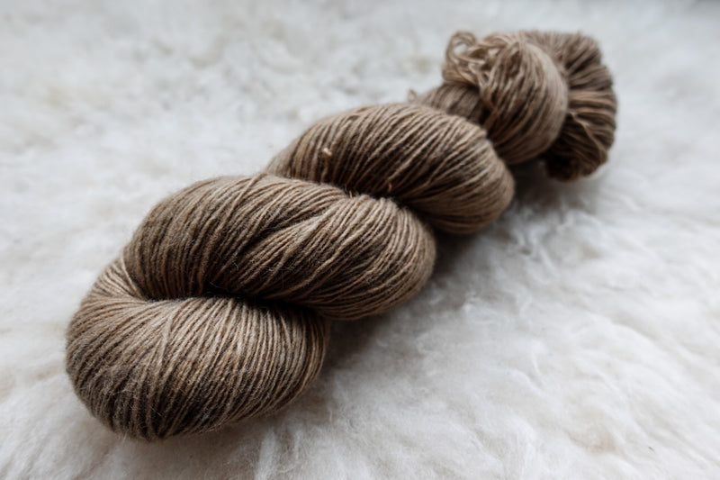 A fingering weight skein of natural fiber yarn has been hand dyed a light brown. It lays on a wool background.