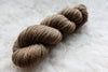 A fingering weight skein of natural fiber yarn has been hand dyed a light brown. It lays on a wool background.