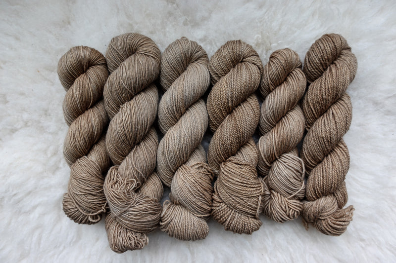 Six skeins of yarn, all different bases, have been hand dyed a light beige. They lay on a sheepskin rug.