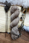A skein of worsted weight yarn has been dyed a light beige. It lays on a tabletop next to dried flowers and bark.