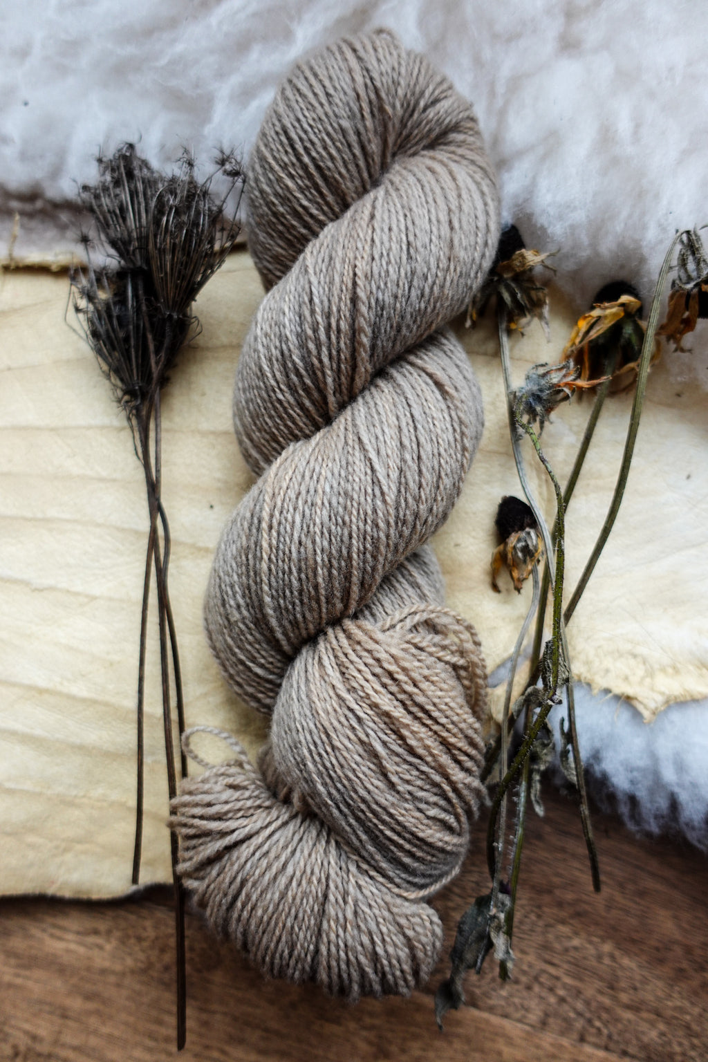 A skein of sport weight yarn has been dyed a light beige. It lays on a tabletop next to dried flowers and bark.
