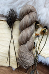 A skein of fingering weight sock yarn has been dyed a light beige. It lays on a tabletop next to dried flowers and bark.