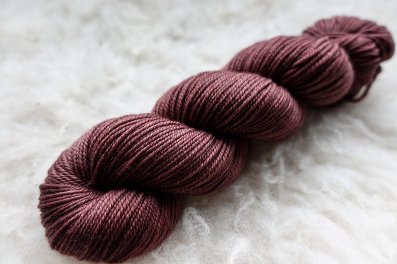 A natural fiber skein of hand dyed, reddish pink yarn lays on sheepskin.