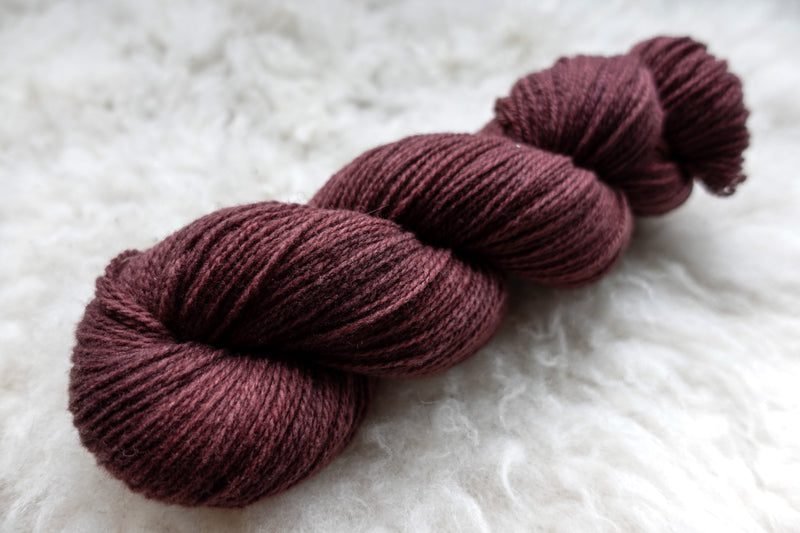 A sport weight skein of natural fiber yarn has been hand dyed a reddish pink. It lays on a wool background.