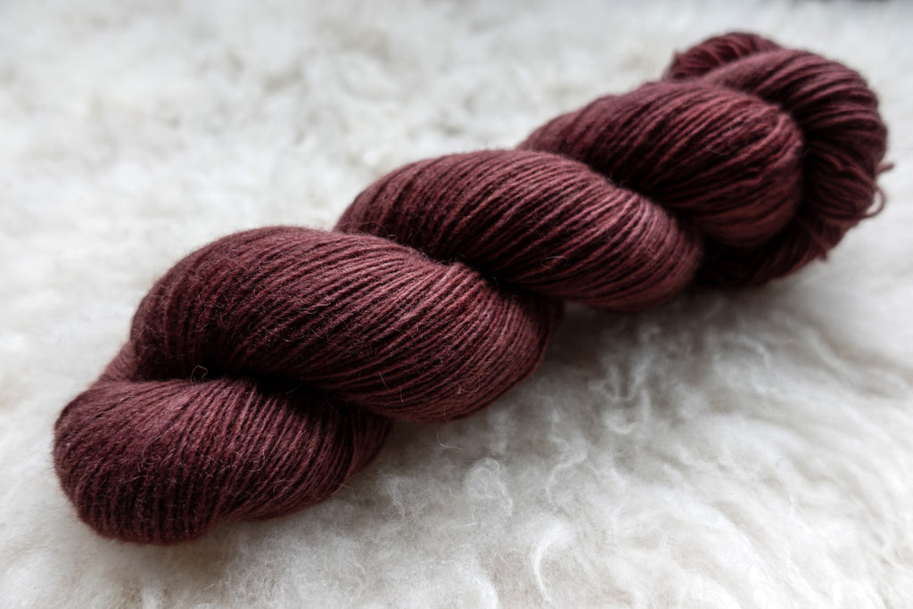 A single ply skein of natural fiber yarn has been hand dyed a reddish pink. It lays on a sheepskin rug.