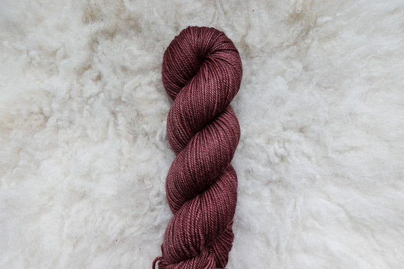 Pictured from above, a hand dyed skein of reddish pink yarn lays on a sheepskin rug.