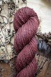 A worsted weight skein of natural fiber yarn has been hand dyed a reddish pink. It lays on a tabletop next to dried flowers.